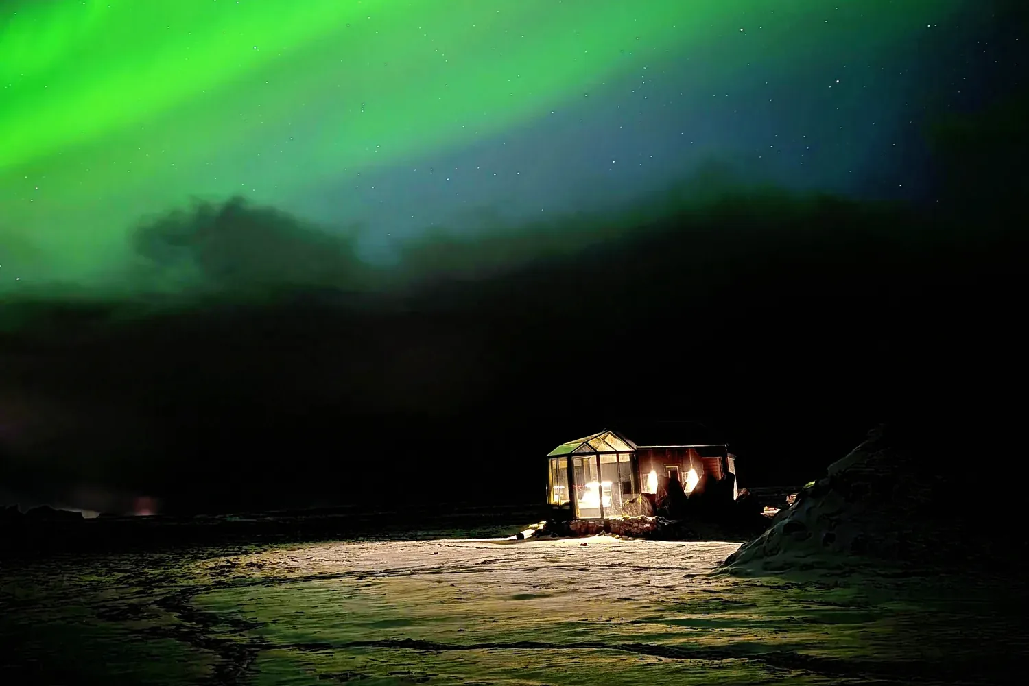 Green northern lights over a lit Aurora Glass Cabin in the snow at Ormskot farm, South Iceland
